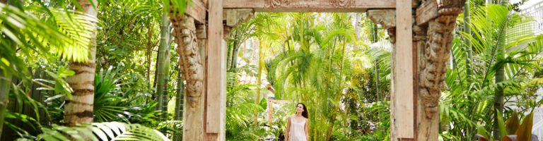 woman walking down a pathway and toward a wooden arch, surrounded by lush tropical greenery